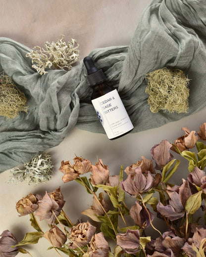 FreeWildShe Cedar & Sage Bitters lay on green fabric surrounded by dried flowers & lichen.