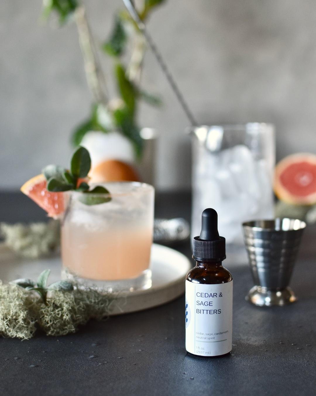 FreeWildShe Cedar & Sage Bitters sit in front of a vibrant pink cocktail with cocktail tools & grapefruit around it.