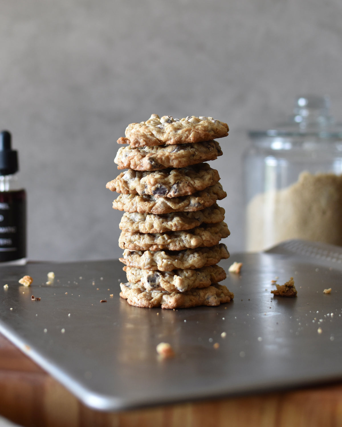 A stack of chocolate chip cookies made with FreeWildShe Barrel Aged Aromatic Bitters sit on a baking tray. A bottle of bitters and glass canister of sugar sit in the background.