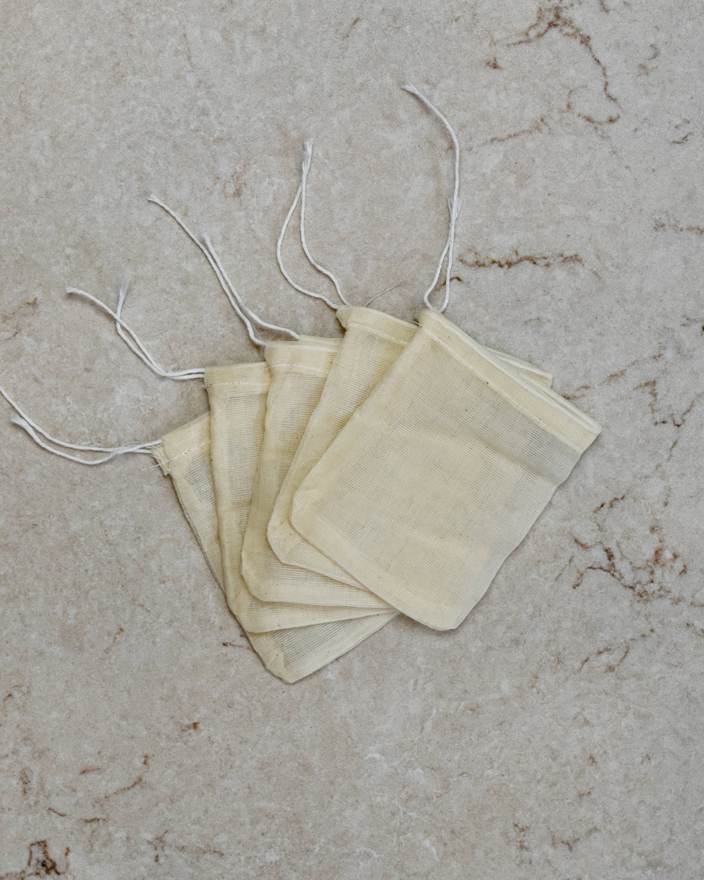 Five FreeWildShe Reusable Cotton Tea Bags lay spread out in the center on a cream colored countertop.