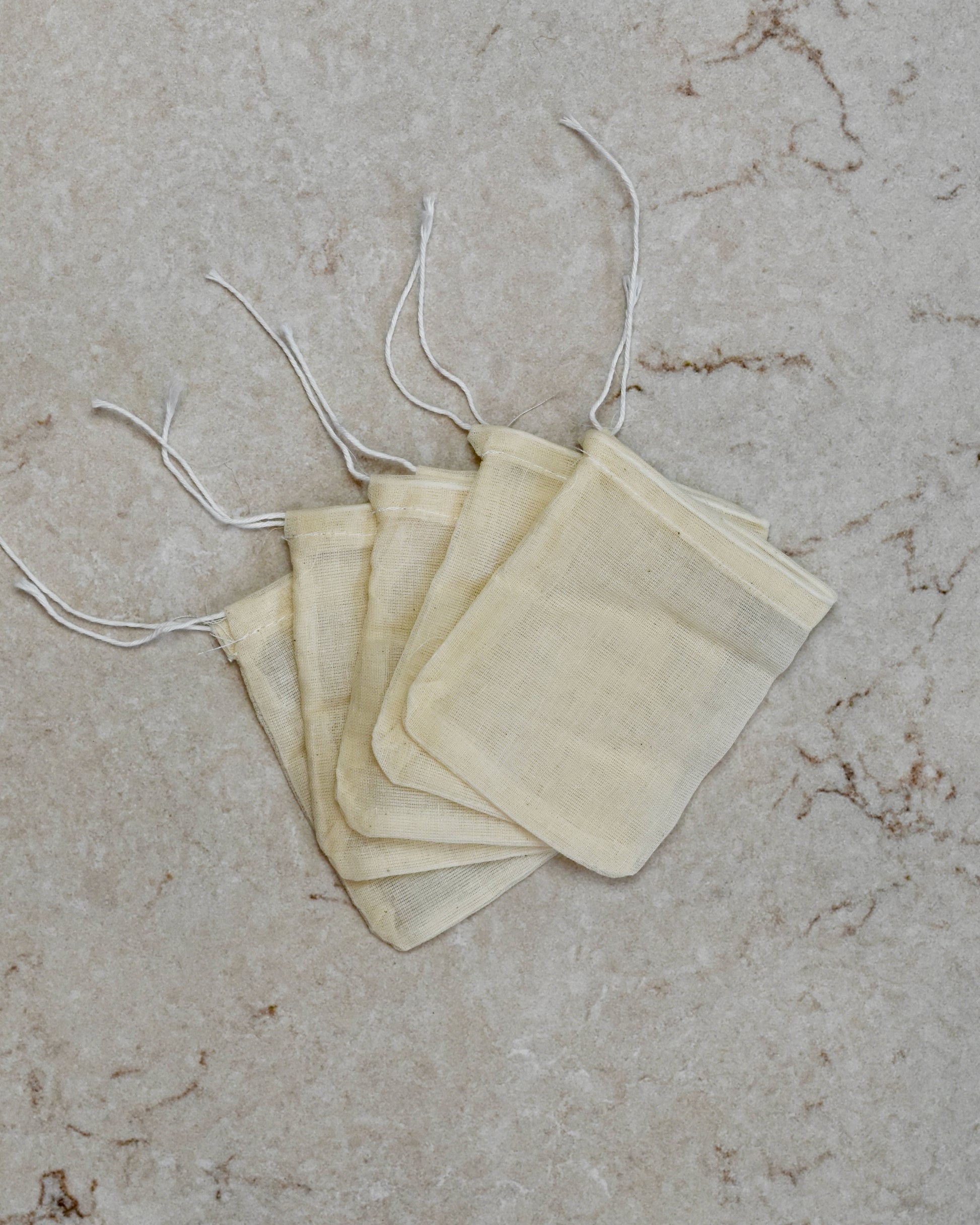 Five FreeWildShe Reusable Cotton Tea Bags lay spread out in the center on a cream colored countertop.