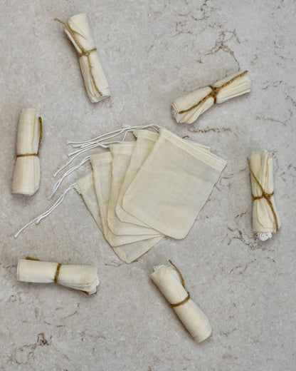 Five FreeWildShe Reusable Cotton Tea Bags lay spread out in the center on a cream colored countertop. Rolled bundles wrapped in twine lay scattered around.