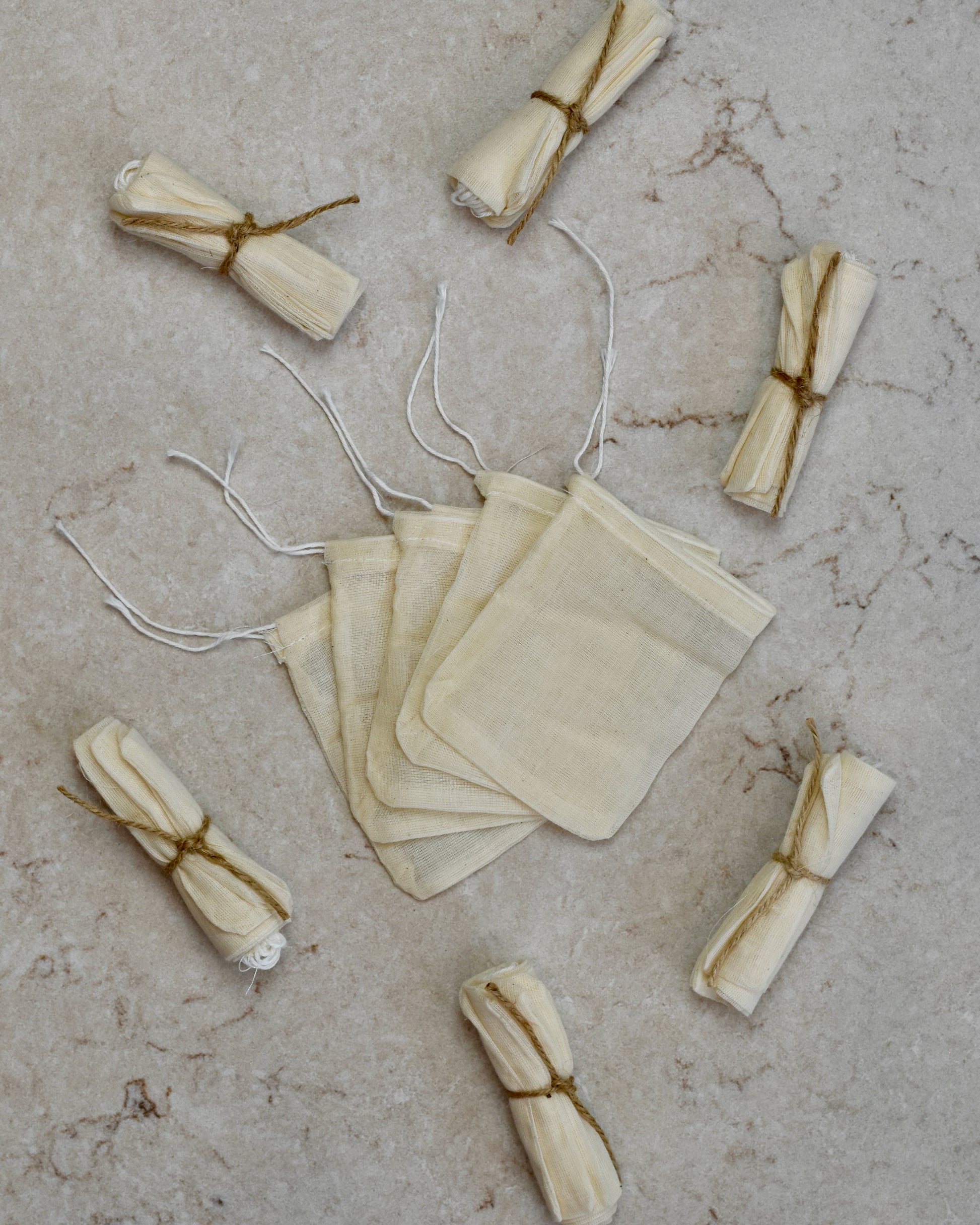 Five FreeWildShe Reusable Cotton Tea Bags lay spread out in the center on a cream colored countertop. Rolled bundles wrapped in twine lay scattered around.