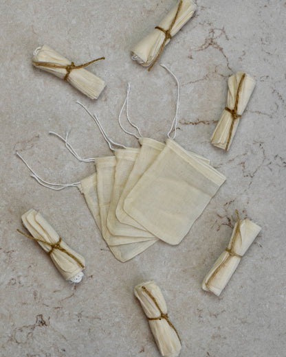 Five FreeWildShe Reusable Cotton Tea Bags lay spread out in the center on a cream colored countertop. Rolled bundles wrapped in twine lay scattered around.