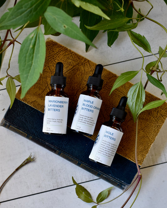 FreeWildShe Marionberry Lavender Bitters, Maple Blood Orange Bitters, & Mole Bitters lay on a book on a white table. They are surrounded by a green, leafy vine.