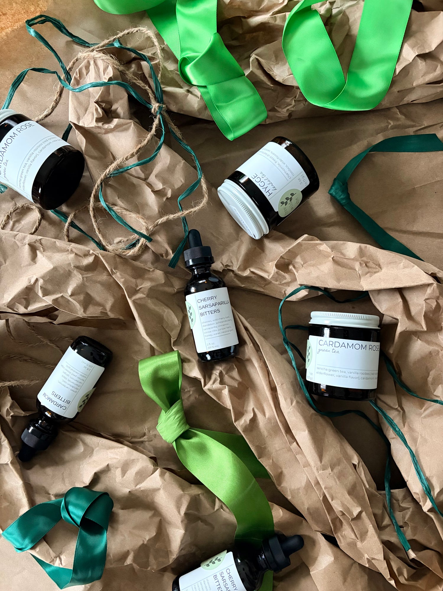 An overhead flat-lay of several small cosmetic or fragrance bottles and jars with white labels, arranged on crumpled brown paper with bright green ribbons and twine.