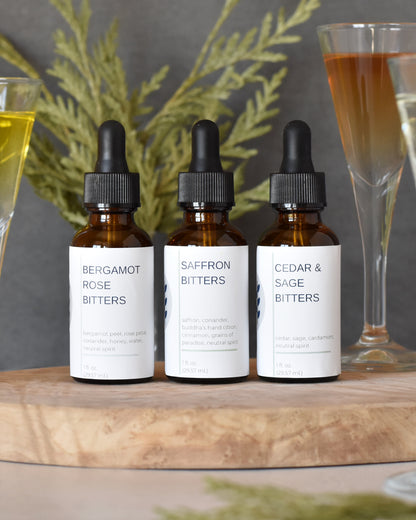 A close up view of FreeWildShe Limited Bitters Collection sitting on a olive wood serving platter. The flavors include: Bergamot Rose Bitters, Saffron Bitters, and Cedar & Sage Bitters. Cocktail glasses with colorful liquid in them and cedar needles are scattered around.