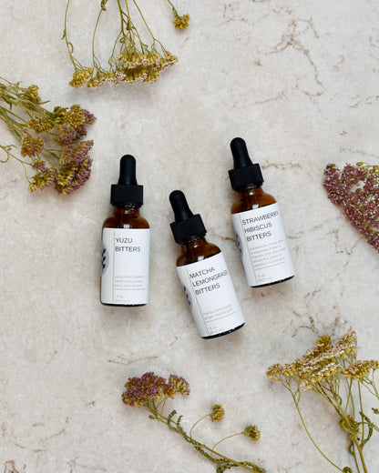 FreeWildShe Limited Bitters Collection - Yuzu Bitters, Matcha Lemongrass Bitters, Strawberry Hibiscus Bitters - lay on a cream colored countertop. Dried flowers are scattered around.