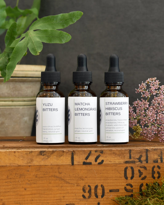 A close up view of FreeWildShe Limited Bitters Collection - Yuzu Bitters, Matcha Lemongrass Bitters, Strawberry Hibiscus Bitters - sit on a wooden box. A plant and dried flower sit in the background.