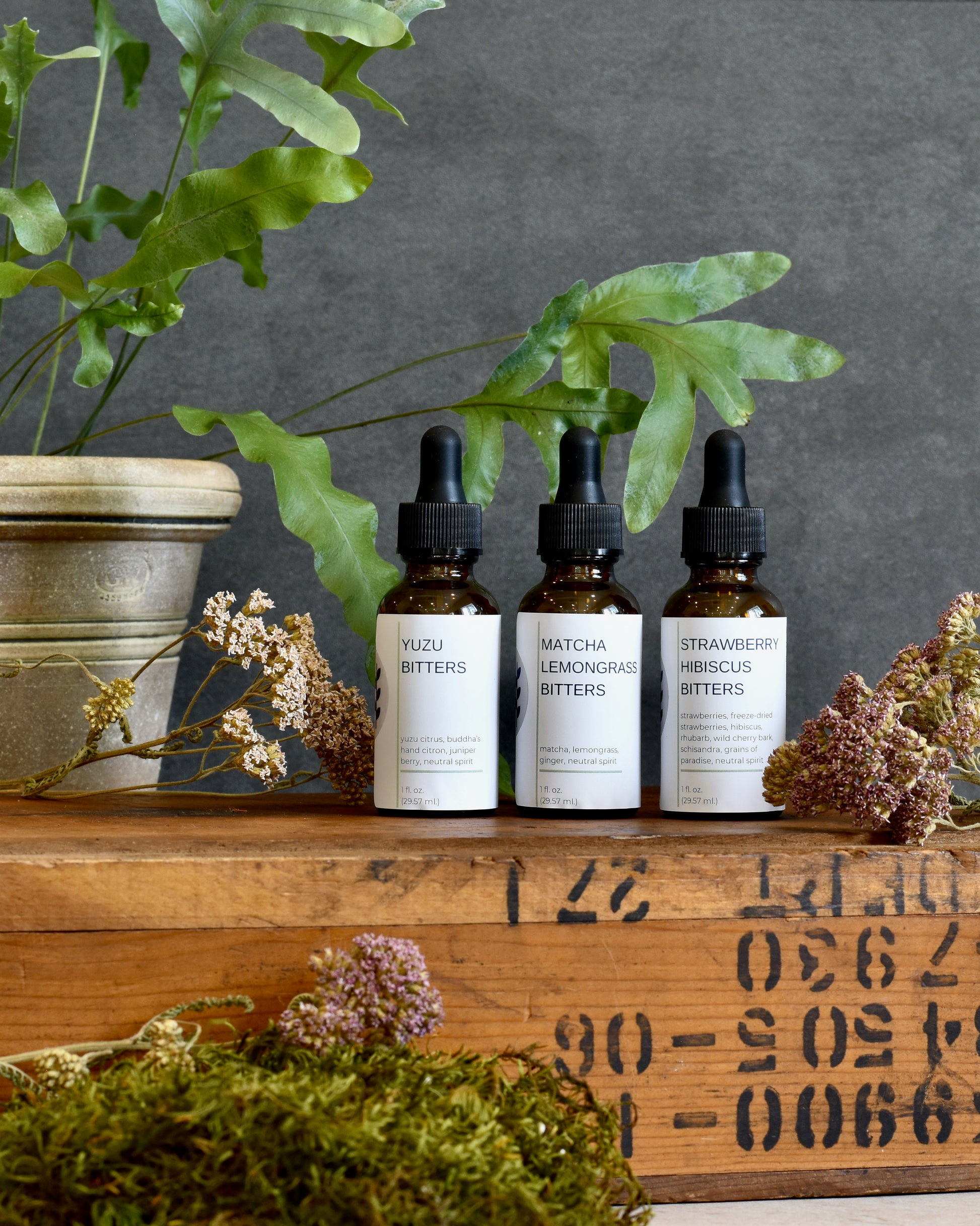 FreeWildShe Limited Bitters Collection - Yuzu Bitters, Matcha Lemongrass Bitters, Strawberry Hibiscus Bitters - sit on a wooden box. A plant sits in the background. Dried flowers are scattered around.
