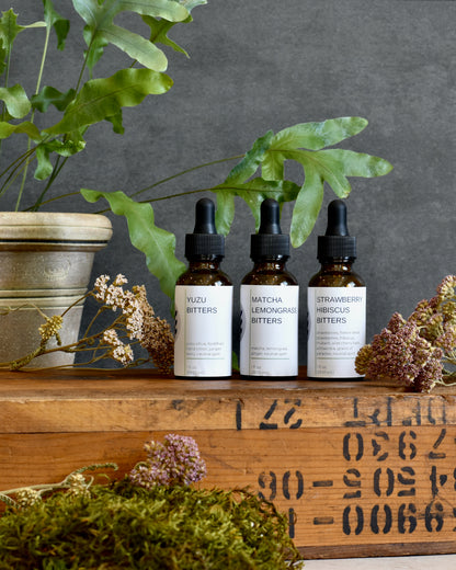 FreeWildShe Limited Bitters Collection - Yuzu Bitters, Matcha Lemongrass Bitters, Strawberry Hibiscus Bitters - sit on a wooden box. A plant sits in the background. Dried flowers are scattered around.