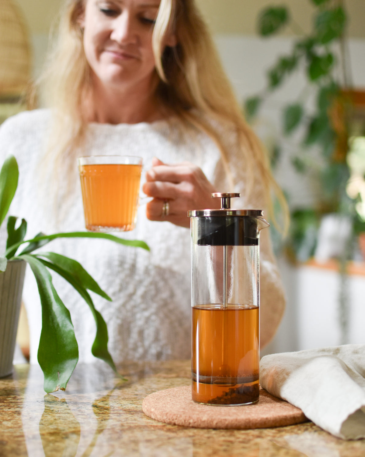 A woman in a white sweater holds a glass of amber tea while standing behind a countertop with a glass French press filled with tea and a cork coaster. Green plants and a warm indoor setting are visible in the background.
