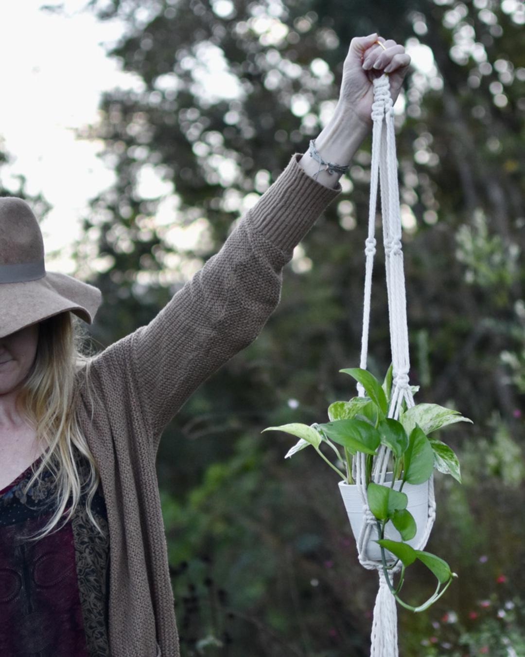 A woman with a hat over her eyes holds a FreeWildShe Ayla Macrame Plant Hanger up in the air. There is a pothos plant in the planter.