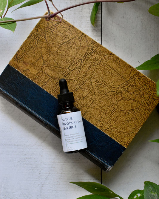 FreeWildShe Maple Blood Orange Bitters lay on an old book on a white, wooden table. A vine and leaves frame the photo.