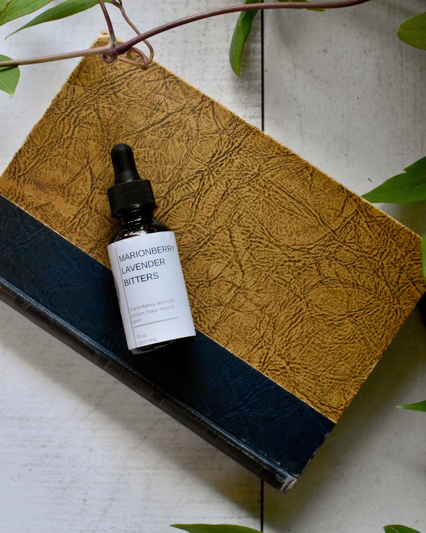 FreeWildShe Marionberry Lavender Bitters lay on an old book on a white, wooden table. A vine and leaves frame the photo.