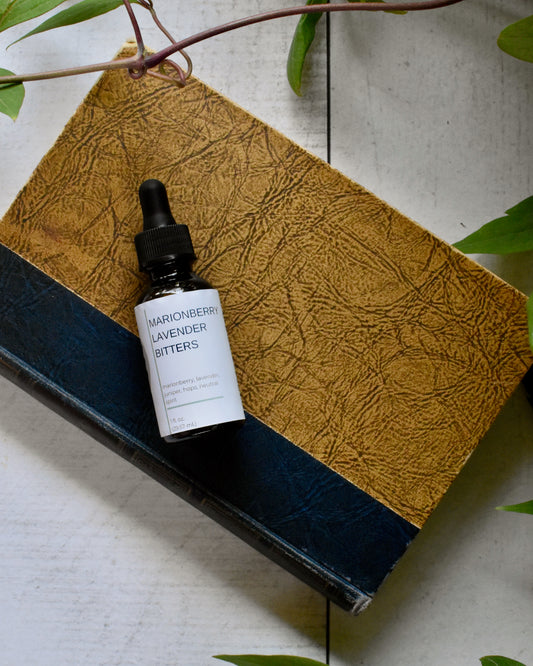 FreeWildShe Marionberry Lavender Bitters lay on an old book on a white, wooden table. A vine and leaves frame the photo.