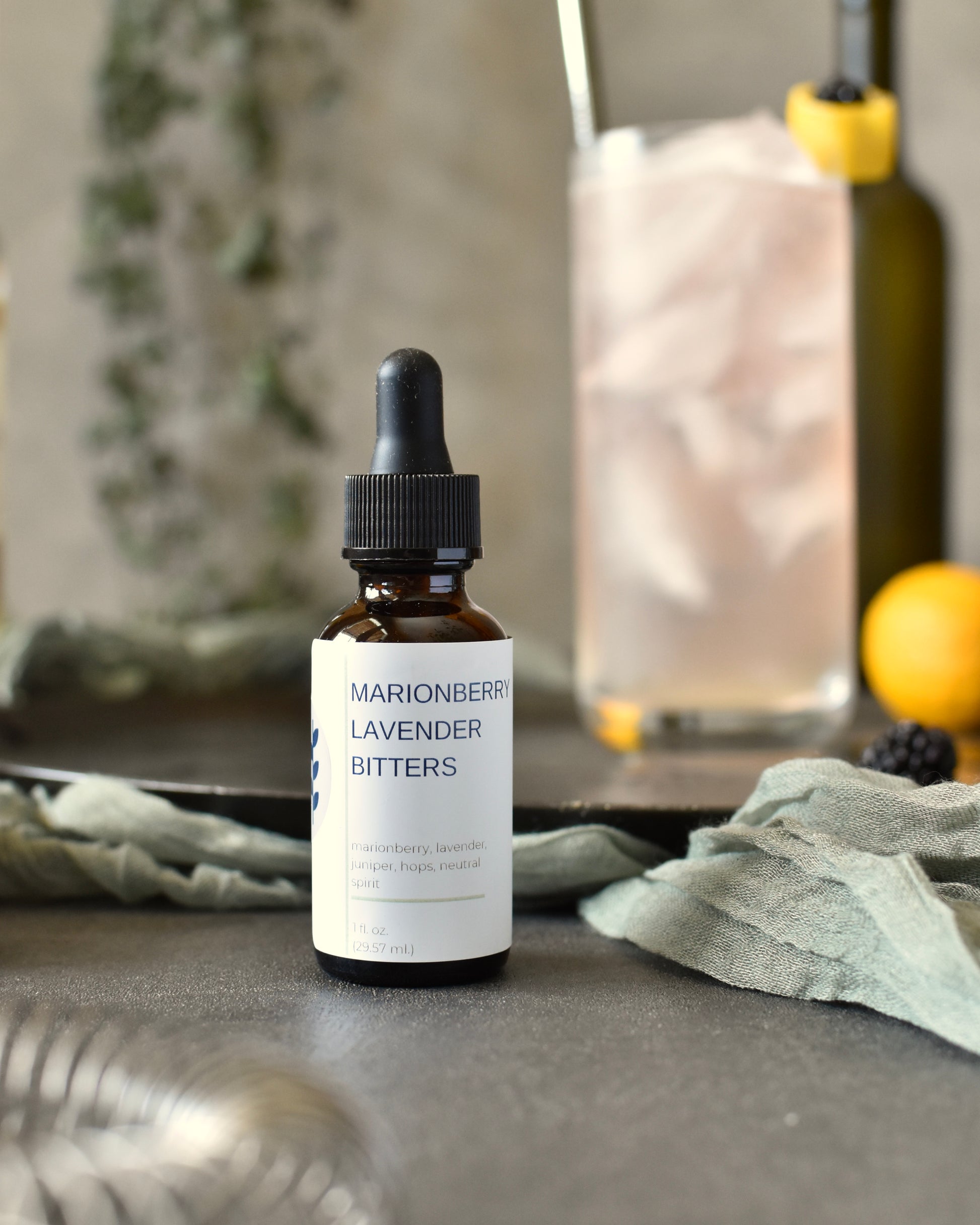 FreeWildShe Marionberry Lavender Bitters sit in front of a Lavender Haze Gin Cocktail. Bar tools, ingredients, and a houseplant are in the background.