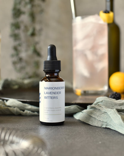 FreeWildShe Marionberry Lavender Bitters sit in front of a Lavender Haze Gin Cocktail. Bar tools, ingredients, and a houseplant are in the background.