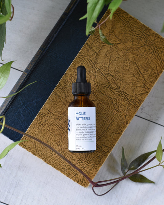 FreeWildShe Mole Bitters lay on an old book on a white, wooden table. A vine and leaves frame the photo.