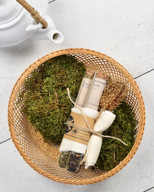 FreeWildShe Mother's Mix loose leaf tea gift bundle lays inside a basked lined with moss. The basket sits on a white wooden table with a white ceramic tea pot next to it. The bundle includes: Calm herbal tea, Madame Noir black tea, and Hygge herbal tea, along with 5 reusable cotton tea bags. They are bundled together with twine and a dried flower.