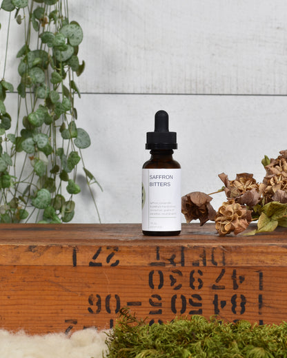 FreeWildShe Saffron Bitters sit on a wooden box. Dried flowers & moss sit in front of the box and a houseplant hangs behind.