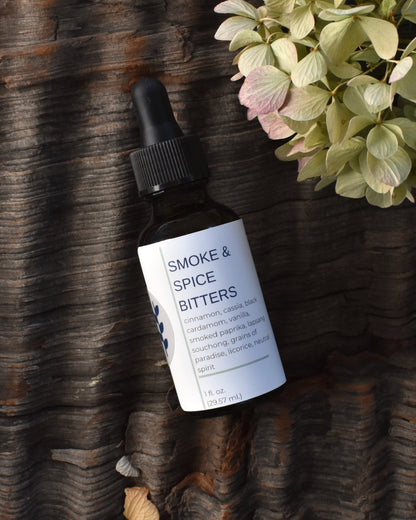 FreeWildShe Smoke & Spice Bitters lay on a textured piece of bark surrounded by dried hydrangea blooms.
