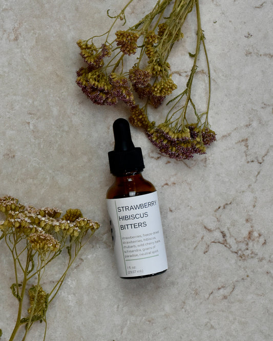FreeWildShe Strawberry Hibiscus Bitters lay on a cream colored countertop. Dried flowers are scattered around.