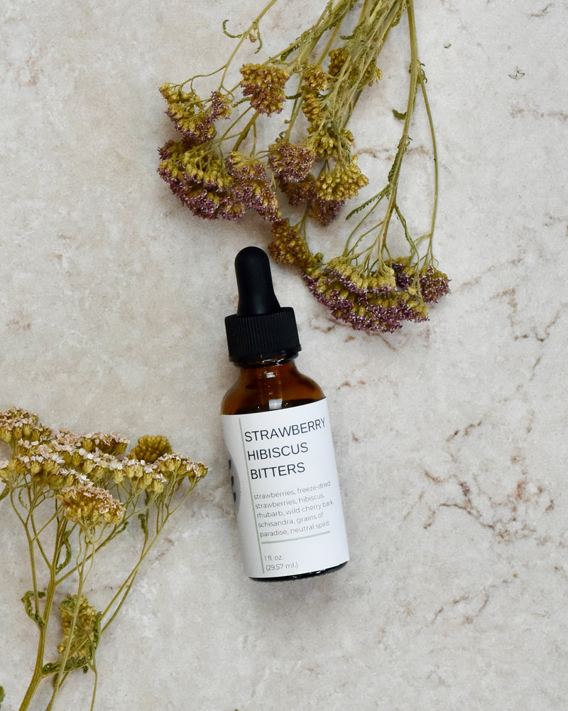 FreeWildShe Strawberry Hibiscus Bitters lay on a cream colored countertop. Dried flowers are scattered around.