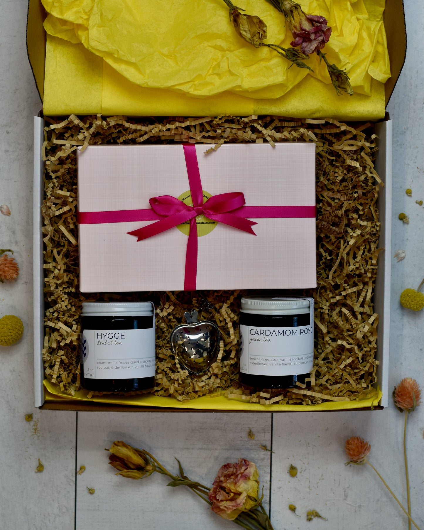 FreeWildShe Mother's Day Bundle is a white box with yellow tissue and kraft crinkle filling the box. Hygge Herbal Tea and Cardamom Rose Green Tea sit in the lower portion. A heart shaped infuser sits between them. A pink box of Suzy's chocolates wrapped in pink ribbon sits in the upper portion.