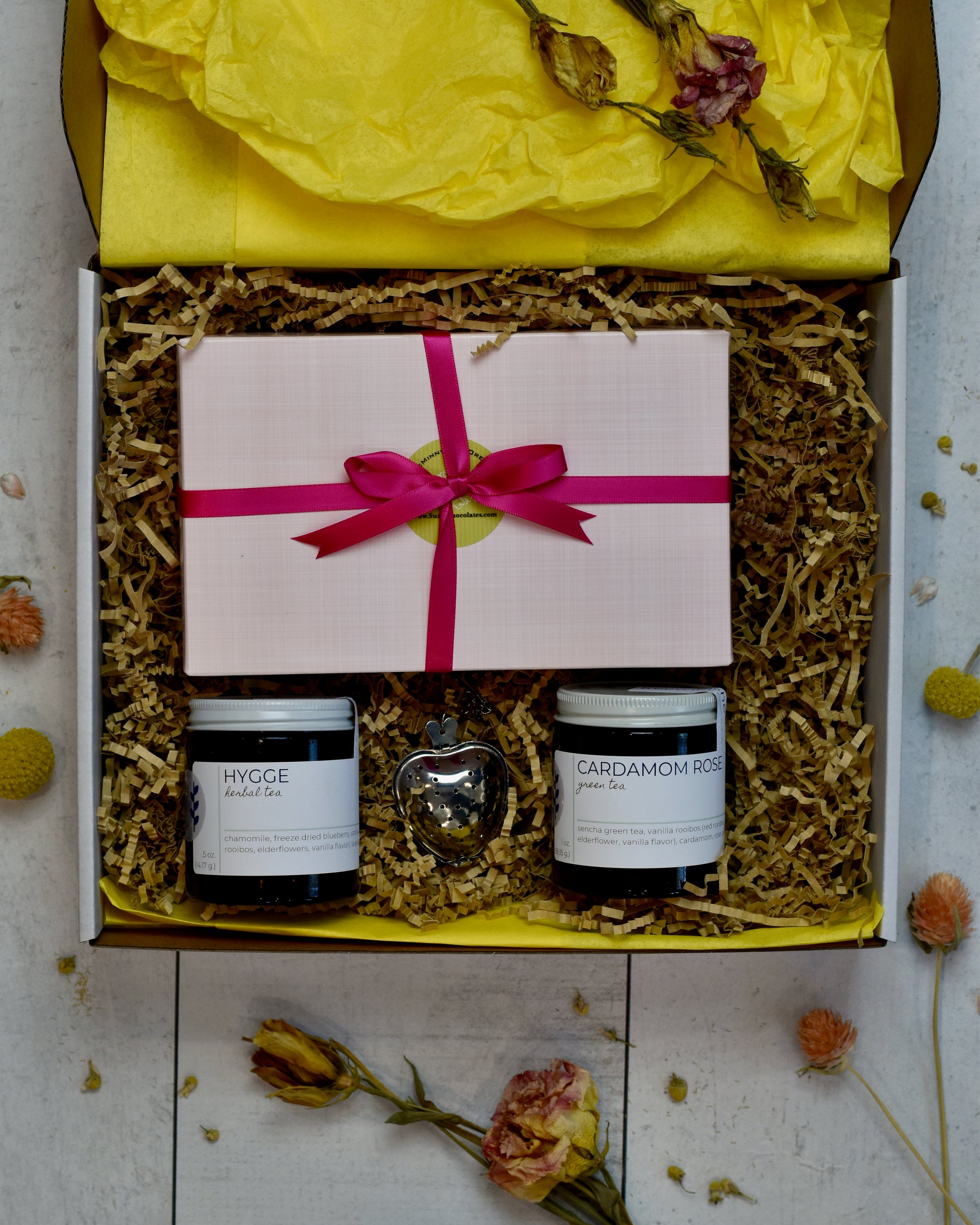 FreeWildShe Mother's Day Bundle is a white box with yellow tissue and kraft crinkle filling the box. Hygge Herbal Tea and Cardamom Rose Green Tea sit in the lower portion. A heart shaped infuser sits between them. A pink box of Suzy's chocolates wrapped in pink ribbon sits in the upper portion.