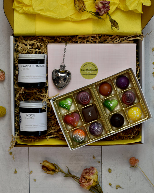 FreeWildShe Mother's Day Bundle is a white box with yellow tissue and kraft crinkle filling the box. Hygge Herbal Tea and Cardamom Rose Green Tea sit on the left side. A heart shaped infuser sits on the pink lid of the chocolate box. Suzy's Chocolates 12-piece box sits open with brightly colored treats of various shapes.