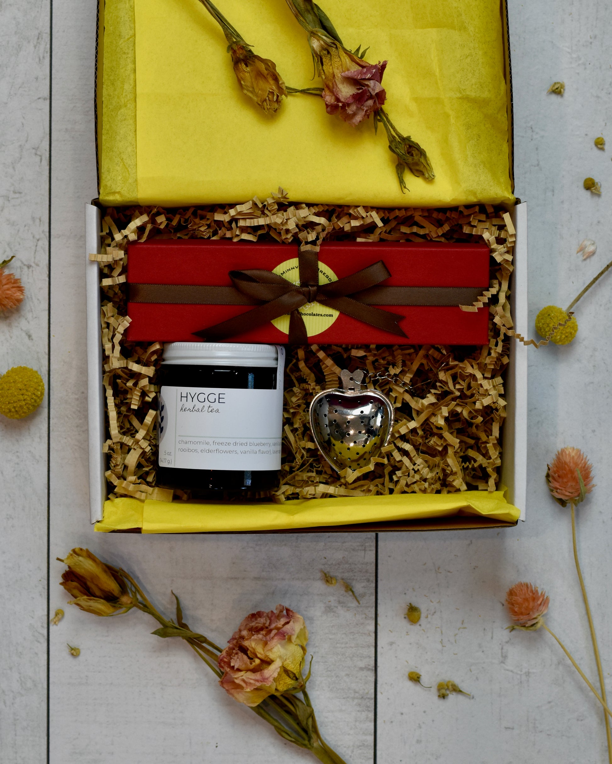 FreeWildShe Mother's Day Bundle is a white box with yellow tissue and kraft crinkle filling the box. Hygge Herbal Tea and a heart shaped infuser sit in the lower portion. Above them, a red box of Suzy's chocolates wrapped in brown ribbon. 