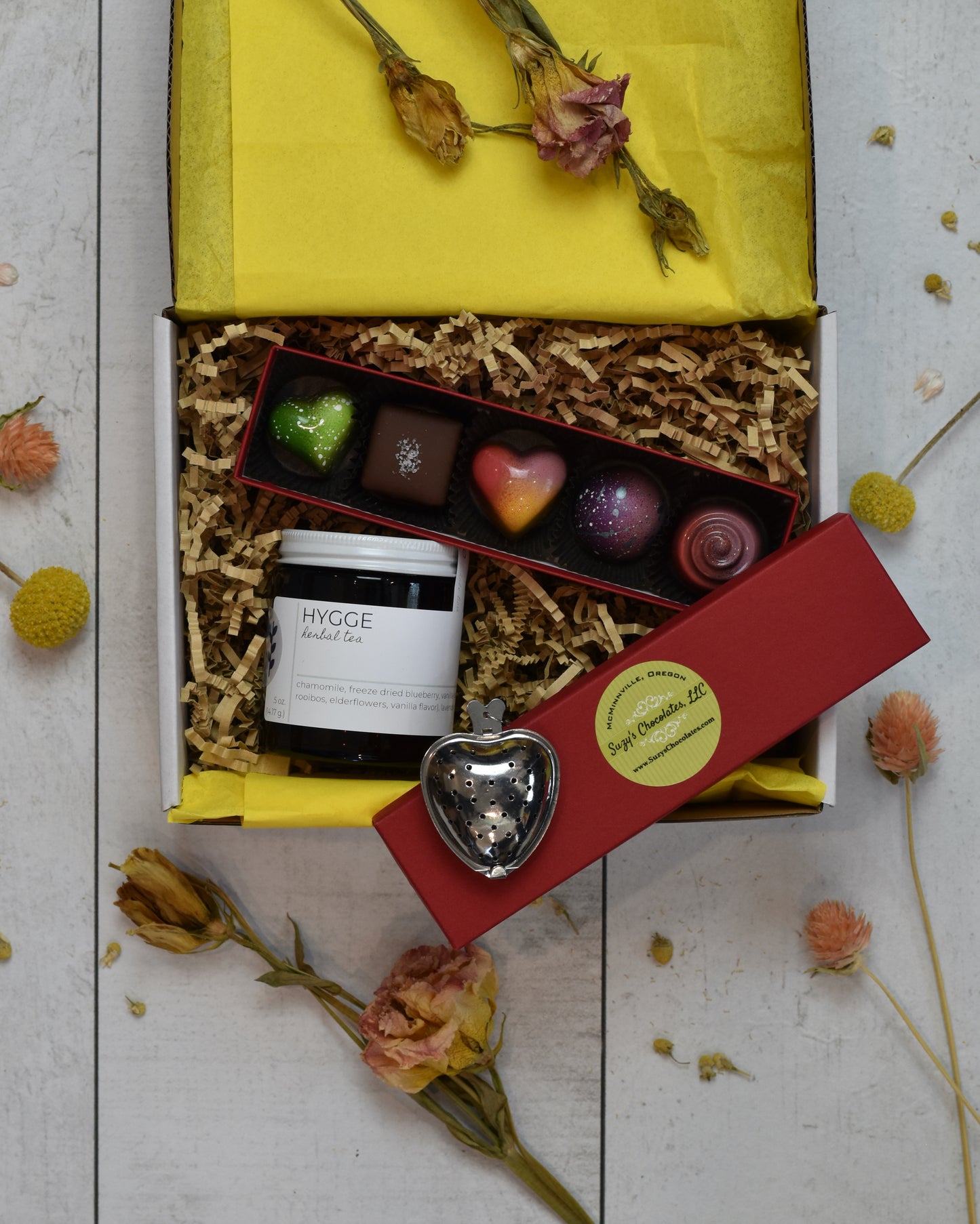 FreeWildShe Mother's Day Bundle is a white box with yellow tissue and kraft crinkle filling the box. Hygge Herbal Tea sits in the lower left corner. A heart shaped infuser sits on the red lid of the chocolate box. Suzy's Chocolates 5-piece box sits open with brightly colored treats of various shapes.