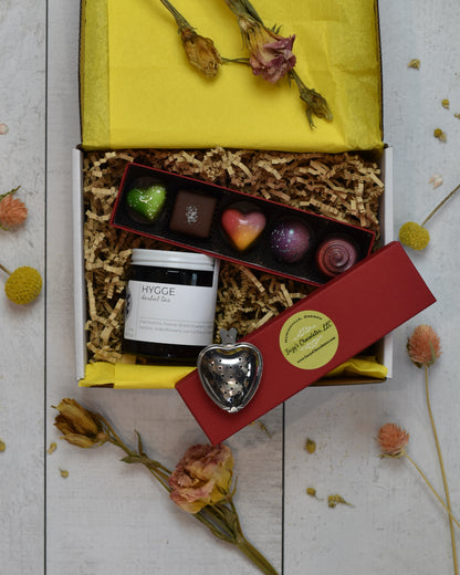 FreeWildShe Mother's Day Bundle is a white box with yellow tissue and kraft crinkle filling the box. Hygge Herbal Tea sits in the lower left corner. A heart shaped infuser sits on the red lid of the chocolate box. Suzy's Chocolates 5-piece box sits open with brightly colored treats of various shapes.