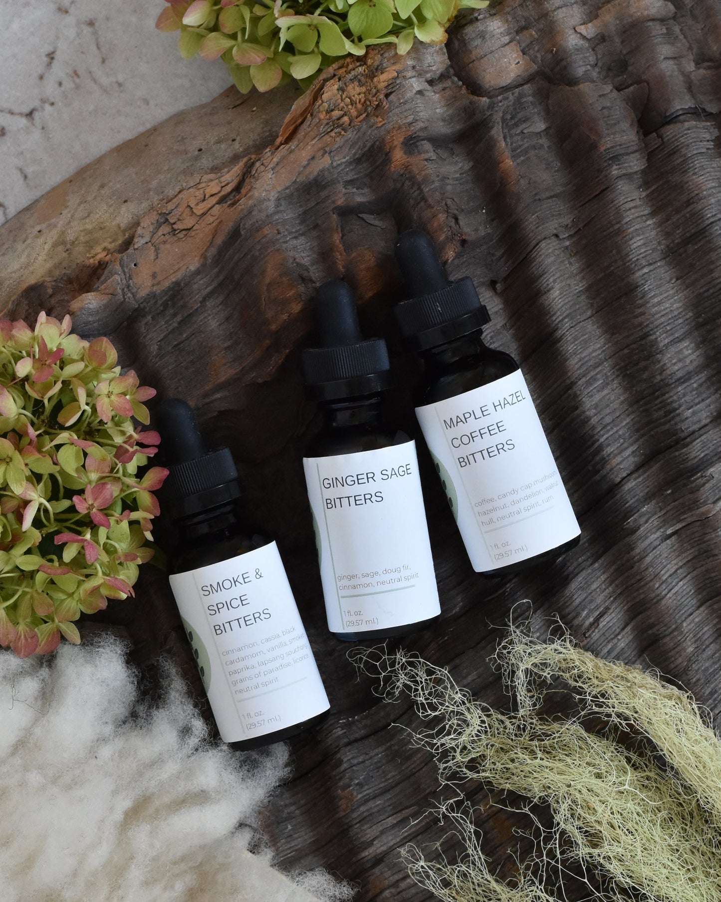 The FreeWildShe Limited Bitters Collection lays on a piece of textured bark surrounded by lichen, hydrangea blooms, and a creamy white sheepskin. From left to right, this trio includes: Smoke & Spice Bitters, Ginger Sage Bitters, and Maple Hazel Coffee Bitters.
