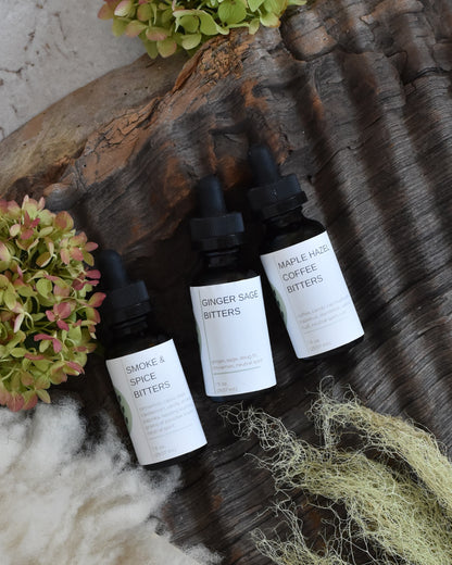The FreeWildShe Limited Bitters Collection lays on a piece of textured bark surrounded by lichen, hydrangea blooms, and a creamy white sheepskin. From left to right, this trio includes: Smoke & Spice Bitters, Ginger Sage Bitters, and Maple Hazel Coffee Bitters.