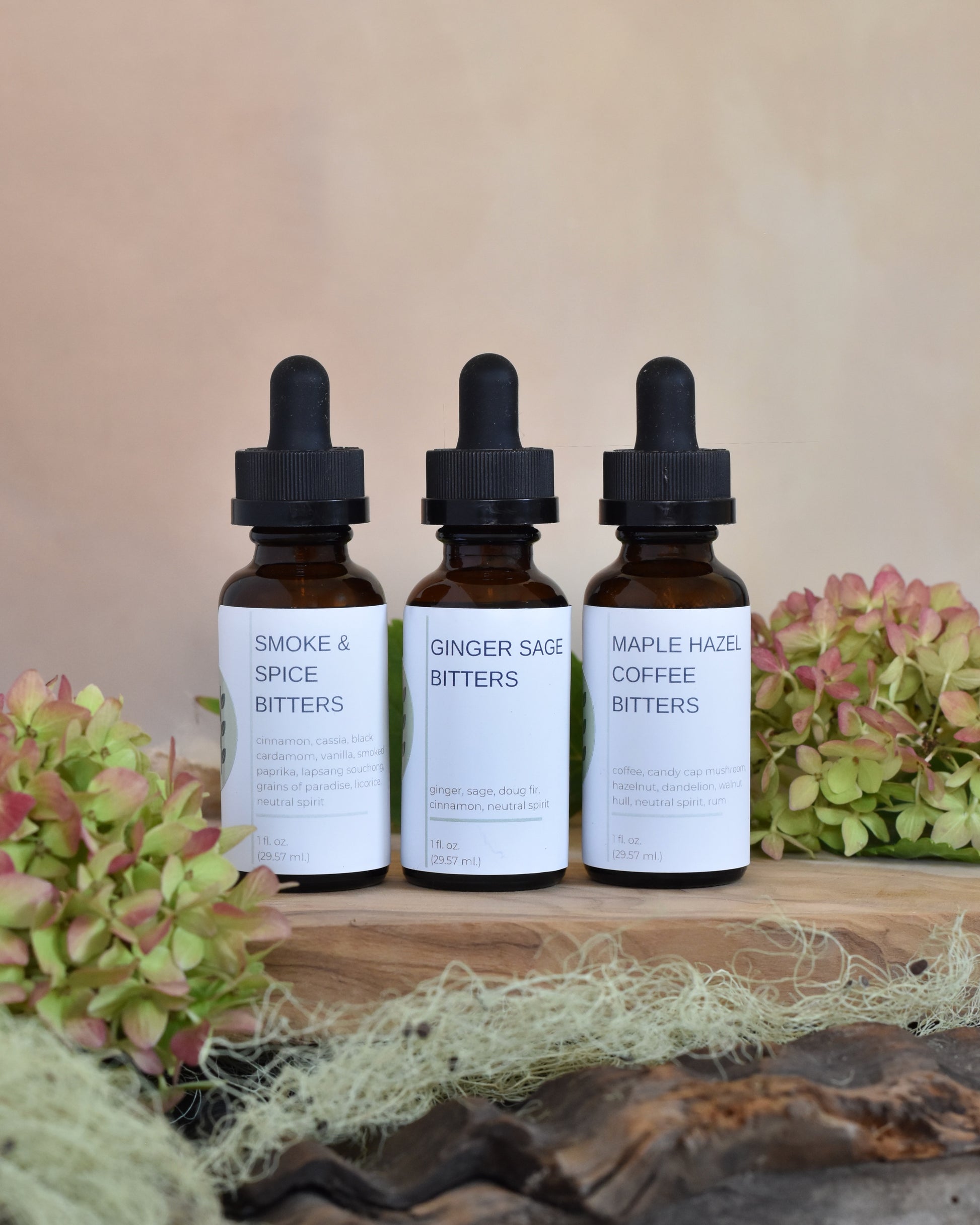 The FreeWildShe Limited Bitters Collection sits on a wood tray surrounded by lichen & hydrangea blooms. From left to right, this trio includes: Smoke & Spice Bitters, Ginger Sage Bitters, and Maple Hazel Coffee Bitters.