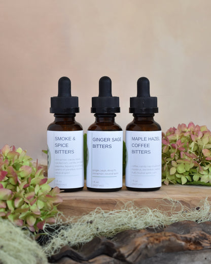 The FreeWildShe Limited Bitters Collection sits on a wood tray surrounded by lichen & hydrangea blooms. From left to right, this trio includes: Smoke & Spice Bitters, Ginger Sage Bitters, and Maple Hazel Coffee Bitters.