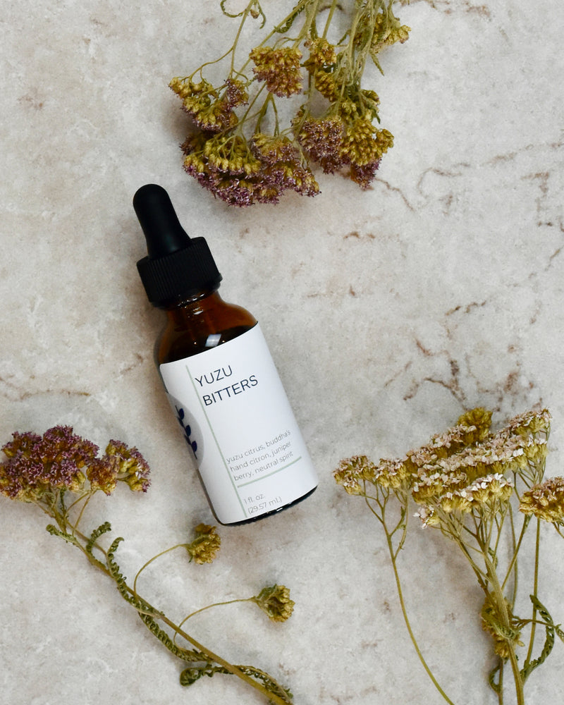 FreeWildShe Yuzu Bitters lay on a cream colored countertop. Dried flowers are scattered around.
