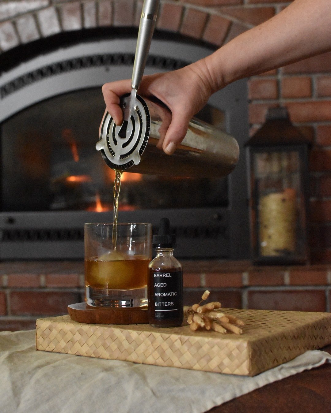 Someone is pouring Hearth Winter Cocktail by FreeWildShe into a glass with ice from a shaker tin. Barrel Aged Aromatic Bitters and dried botanicals sit beside it. There is a fireplace in the background.