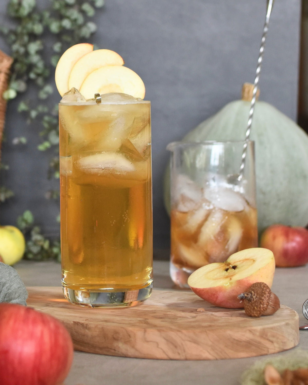 A Honey Pie cocktail by FreeWildShe with Chamomile Vanilla Bitters sits on a wooden platter. Apples, a mixing glass, and a sage green squash sit around the scene.
