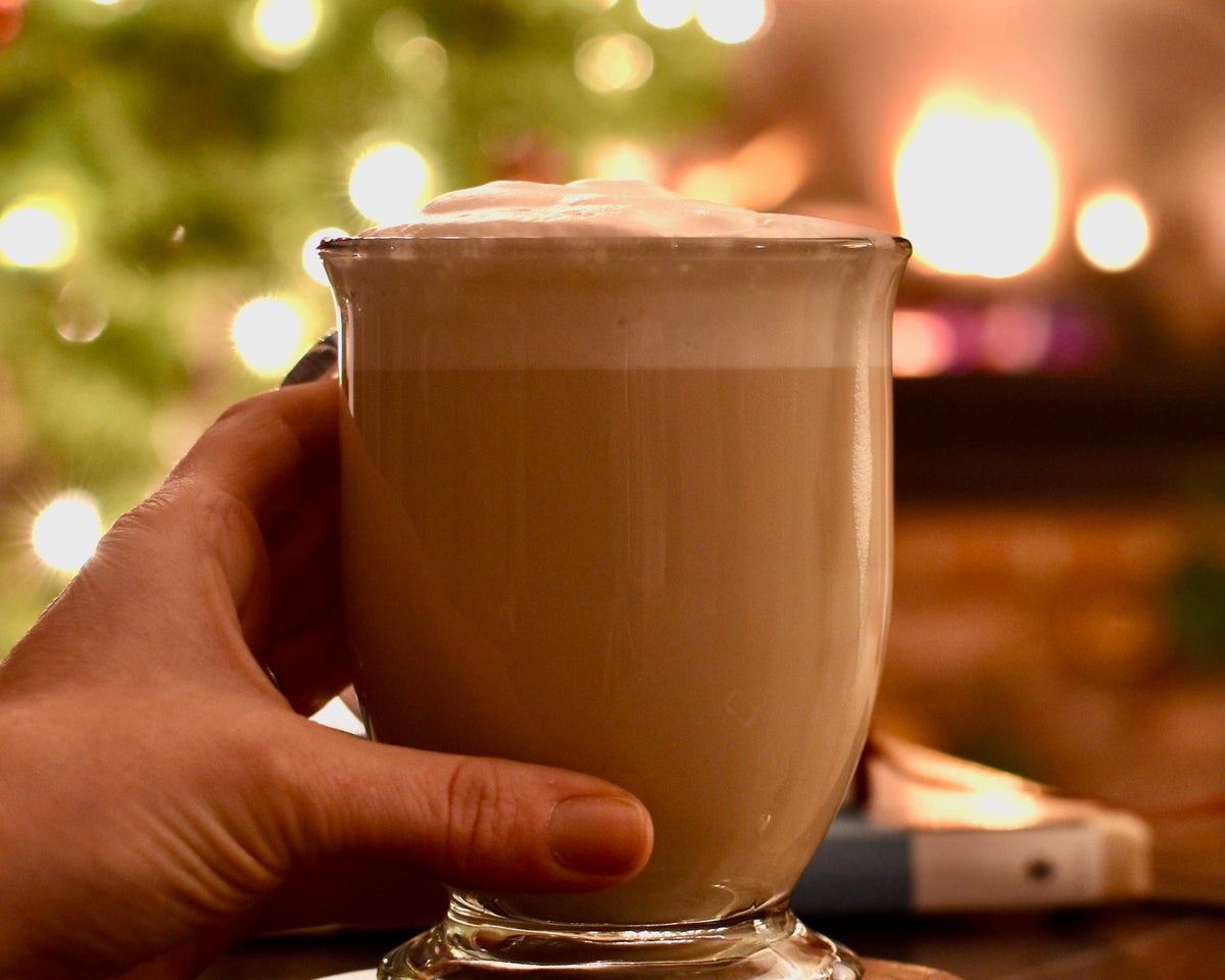 A mug of Long Night's Moon Hygge Herbal Tea sits on a table. A hand wraps around the mug. A Christmas tree & fireplace are blurred in the background.