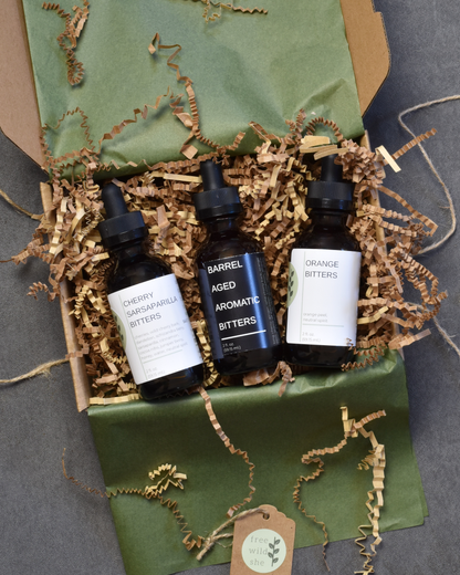 The Old Fashioned Bitters Bundle including Cherry Sarsaparilla, Barrel Aged Aromatic, and Orange Bitters lays a box with olive green tissue and brown crinkle stuffing.