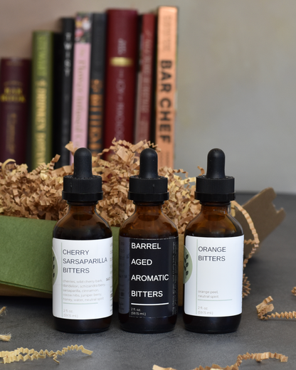 The Old Fashioned Bitters Bundle including Cherry Sarsaparilla, Barrel Aged Aromatic, and Orange Bitters stands in front of a box with olive green tissue and brown crinkle stuffing. A stack of cocktail books stands in the background.