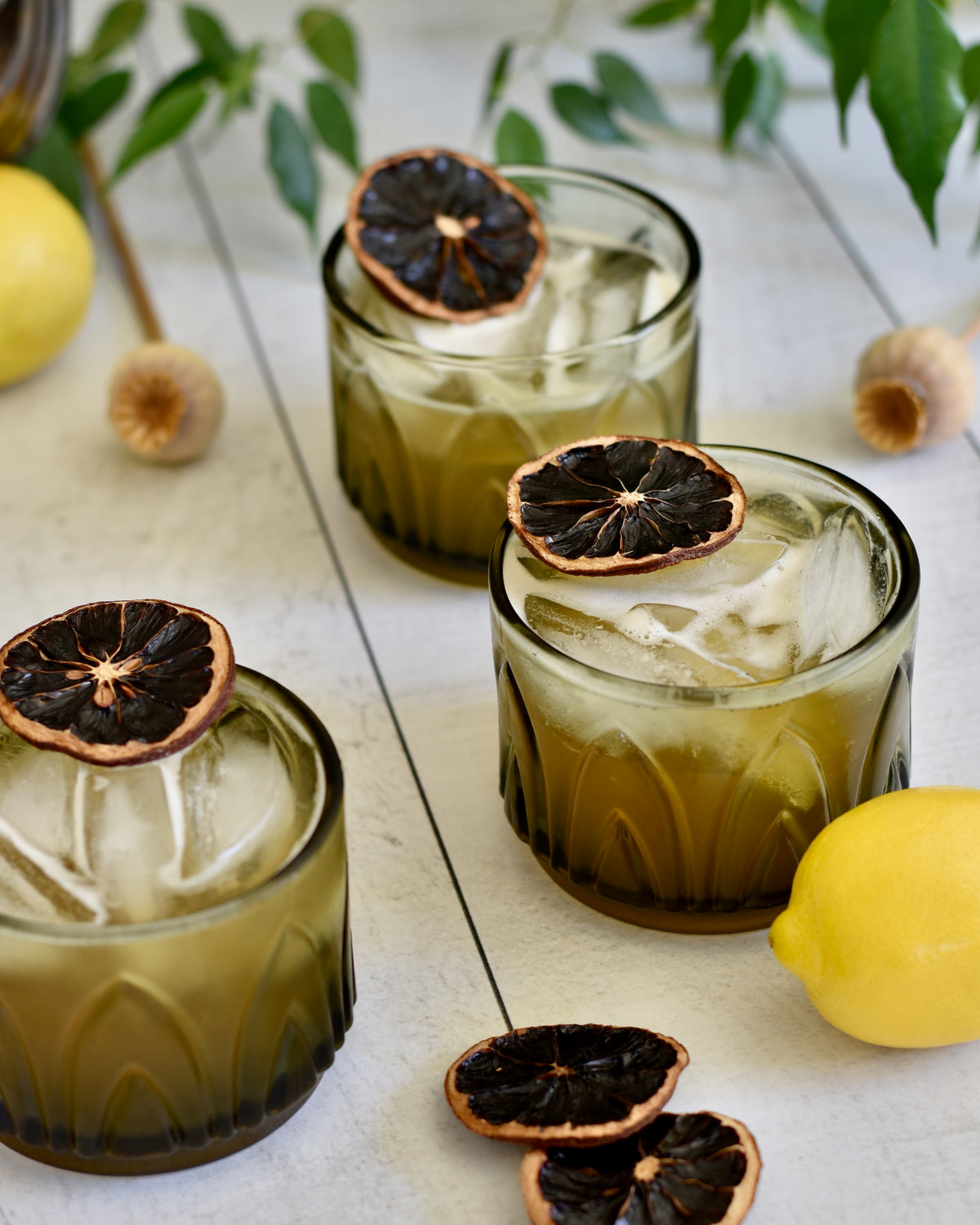 Three Palmer In Bloom cocktails by FreeWildShe sit on a white wooden table. They are surrounded (and garnished) with dried lemon wheels. Lemons, dried seed pods, and a plant surround the scene.