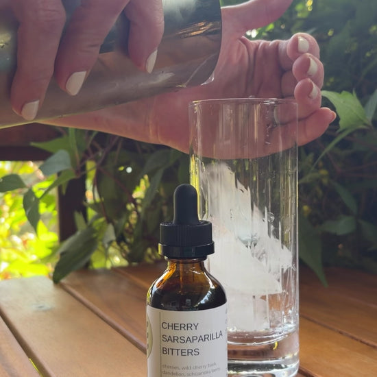 A video of someone making a Toasted Coco-Cherry Tonic by FreeWildShe. A bottle of Cherry Sarsaparilla Bitters sit in front of a glass. They both sit on a wooden table outside with a green vine in the background.