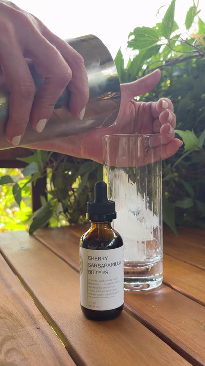 A video of someone making a Toasted Coco-Cherry Tonic by FreeWildShe. A bottle of Cherry Sarsaparilla Bitters sit in front of a glass. They both sit on a wooden table outside with a green vine in the background.