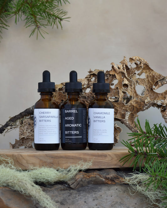 The whiskey lover's bitters bundle sits on a wooden tray surrounded by lichen & douglas fir. From left to right, his trio includes: Cherry Sarsaparilla Bitters, Barrel Aged Aromatic Bitters, and Chamomile Vanilla Bitters.