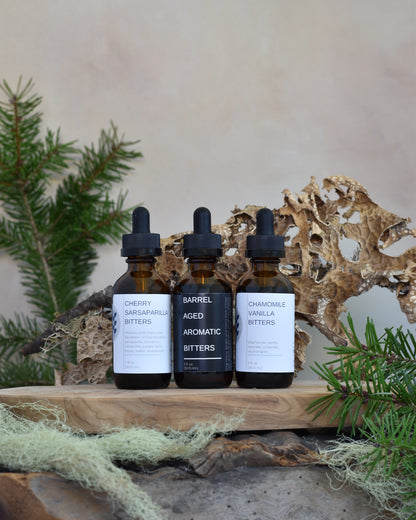 The whiskey lover's bitters bundle sits on a wooden tray surrounded by lichen & douglas fir. From left to right, his trio includes: Cherry Sarsaparilla Bitters, Barrel Aged Aromatic Bitters, and Chamomile Vanilla Bitters.